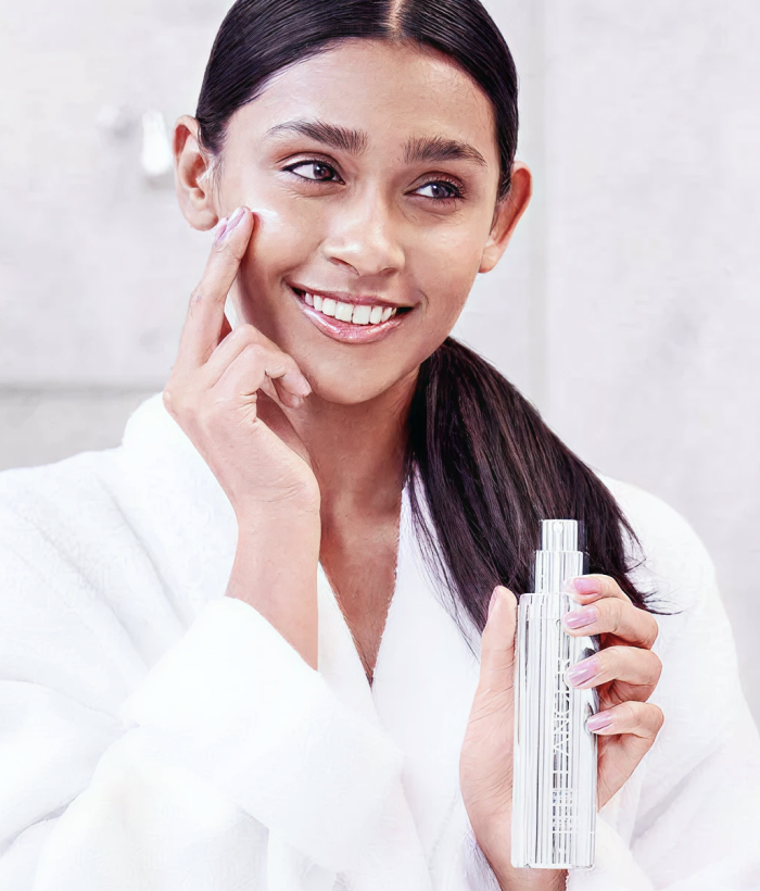Woman applying cream to her face with a skincare product in hand, wearing a white robe.-Lancer Skincare